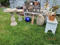 Wide view of garden decor items on lawn including two composite bird baths, blue gazing ball, owl sculptures, vintage watering cans, lanterns, flower pots, and two painted benches