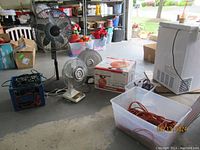 Full overview of lot showing Ivy portable fan, Lasko fan, Presto heater box, extension cords in plastic container, and other storage.