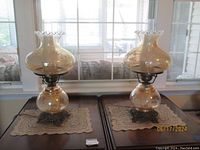 Pair of vintage amber glass Gone with the Wind style hurricane lamps shown on a table with crocheted doilies, detailed with white frost floral patterns and ornate metal bases.