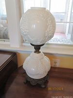 Full view of vintage Victorian double globe milk glass lamp with grapevine pattern on globes, ornate metal base, standing on a table near window.