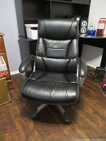 Front view of a black leatherette office chair with padded seat, backrest, and armrests, positioned on a hardwood floor in an office setting.