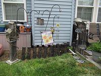 View of tall bird house stand, iron shepherd hooks with hanging ornaments, and solar garden lights on the grass in front of a light blue house wall.