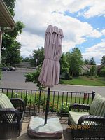 Beige outdoor patio umbrella closed and mounted on base, shown with patio chairs and background of neighborhood street and greenery.
