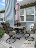 Full view of outdoor dining set featuring a metal framed square table with tiled top, four swivel chairs with brown mesh fabric and beige cushions, beige closed umbrella in center, and outdoor area rug underneath.