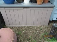 Large beige plastic outdoor storage box with wood panel design shown closed outside on soil.
