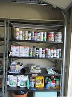 Metal shelving unit with assorted large paint cans, spray paint cans, painting rollers, boxes of light bulbs, garden care sprays and fertilizers on shelves