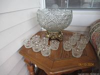 Full view of the cut glass punch bowl on brass stand surrounded by 12 matching cut glass cups arranged on a wooden table.