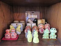 Twelve assorted collectible salt and pepper shaker sets including turkeys, frogs, gingerbread people, and various animal and character shapes, displayed on wood shelf.