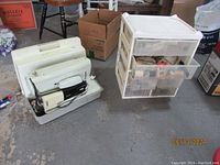 Singer sewing machine in hard plastic case alongside a plastic container with sewing notions on concrete floor, surrounded by boxes and other items.