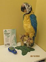 Front view of Squawkers McCaw plush parrot standing on green leafy base with remote control and pamphlet beside it.