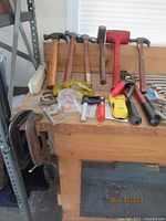Full view of assortment of hand tools on wooden workbench including hammers, clamps, crimp tool, pliers, measuring tape, screwdriver, and safety glasses.