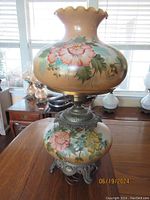 Full view of the 1940s hand painted hurricane lamp showing large floral design on glass globe shade and base with ornate metal stand on wooden table near windows.