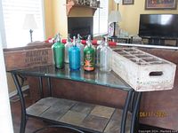 Photo showing nine vintage glass seltzer bottles in green, blue, and clear glass placed on a glass table with two wooden crates behind them. The wooden crate on the left is labeled Raritan Beverage Co. Nixon N.J., and the crate on the right is labeled Pabco - Eagle.