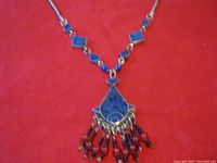 Front view of the pendant necklace showing the blue stone pendant with silver swirl design and attached beaded tassels against a red background.