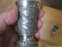 Close-up of pewter cup with detailed relief of a stag and doe in a forest setting, showing the textured base and ornamental details.