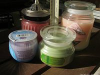 Four scent candles on surface; visible colors pink, cream, red, green, mauve; visible labels showing some scents