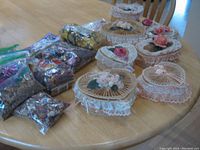 Photo showing eight lidded baskets of oval and heart shapes with lace and artificial flower decorations, arranged on table alongside multiple sealed bags of potpourri and one small bag of waxed pomander.