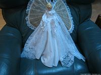 Angel Christmas tree topper sitting on a dark green chair showing full dress and wings from front angle.