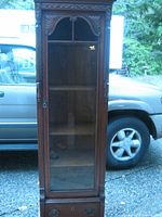 Front view of antique china cabinet showing wooden frame, glass door, carved top detail, and brass top rail.