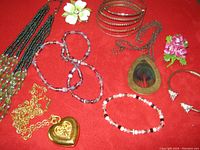 Full view of costume jewellery on red fabric, includes heart locket necklace, peacock feather pendant, flower pins, joined bangles, multiple beaded bracelets and necklaces