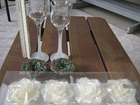 Full view of two glass candle holders, four white rose-shaped floating candles in plastic packaging, and two small decorative green wreaths with red holly berries on a wooden surface.