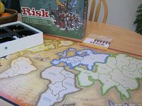 Risk game board laid out on table with territories and colored zone boundaries visible. Box in background with Risk logo and artwork.