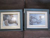 Two framed nature art prints side by side on a couch, one featuring a squirrel and the other a rabbit in snowy landscapes.