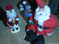 Photo showing two Santa figurines: one plug-in model holding an item and one large stuffed Santa sitting on the floor.