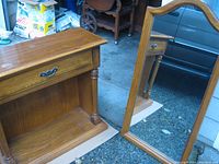 Wooden hallway table with drawer and open lower shelf, shown standing on floor with visible turned side columns and metal handle on drawer. Large wooden framed mirror leaning next to it, showing reflective surface and frame detail.