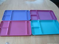 Four plastic TV trays of different pastel colors (blue, pink, purple, turquoise) arranged on a oval wooden table showing their divided compartments.