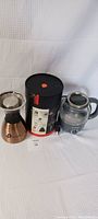Asobu coffee drip maker, packaging tin, and Elite Gourmet glass kettle shown side by side on white background.
