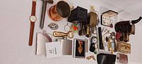 Photo showing assortment of metal banks, flasks, a watch with brown leather strap, a few keychains, and cigar accessories.
