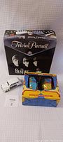 Photo showing the Beatles Trivial Pursuit board game box, two Yellow Submarine drinking glasses in original packaging, and the small white diecast London taxi car placed in front of the box.