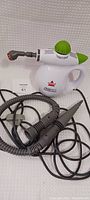 Front and side view of white and green Bissell handheld steam cleaner with attached hoses and nozzle attachments laid on white background.