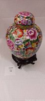 Full view of large Chinese ginger jar on wooden pedestal, showing overall shape, floral painted surface, and lid.
