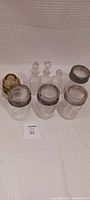 Photo of seven vintage Crown glass containers with metal lids and four small clear glass stopper bottles arranged on white surface.