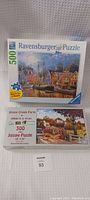 Two vintage jigsaw puzzle boxes: Ravensburger 500-piece 'Coastal Marina Scene' on top and Sunsout 300-piece 'Stone Creek Farm' by William A. S. Kreutz below, both new in box.
