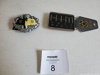 Lot of 2 belt buckles displayed side by side on a white surface. Scarface buckle is silver and yellow with decorative flames and initials. HIM buckle is black with silver text and an attached tag with a gold pentagram logo.