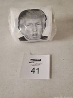 White toilet paper roll with black and white images of Donald Trump's face, packaged in clear plastic, photographed on a white surface with lot number card.