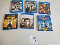 Photo of all six Blu-ray movie cases arranged flat showing front covers including Zombieland, Boondock Saints, Hunger Games, Aviator's Method, Moan, and Happy Feet Two.