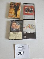 Photo showing 4 music cassette tapes arranged in a square on a plain white surface, with a MaxSold lot number card placed below.