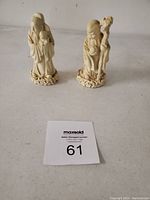 Two ivory-colored statues of Asian figures placed on a flat surface with a lot number card.