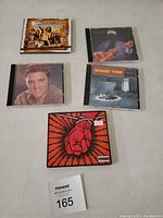 Top view showing five compact discs with varied album covers on a neutral surface, including Elvis Presley and Madder Rose albums.