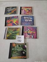 Top-down photo of 7 CD jewel cases arranged on a surface showing their front covers, various music genres represented including dance and karaoke.