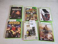 Image showing six Xbox 360 game cases arranged in two rows of three on a white surface. Titles visible include Singularity, Borderlands 2, Prototype, Duke Nukem Forever, Battlefield Bad Company, and Call of Duty Modern Warfare 2.
