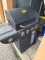 Front and side view of large black gas BBQ grill showing four main burner control knobs, side burner knob, temperature gauge on hood, and orange trim on handles and knobs.