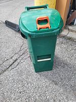 Green compost bin placed outdoors on asphalt, showing front and side, with orange handle on lid and side grip, ventilation slats near base.