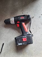 Side profile of a black and red cordless Jobmate drill with an 18.0V battery attached, placed on a concrete/gray surface.