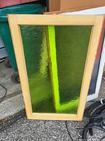 Rectangular window with textured green glass and a light brown wooden frame leaning against a wall near a green recycling bin and some outdoor equipment.