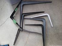 Seven metal L brackets of various sizes and colors arranged on concrete floor.